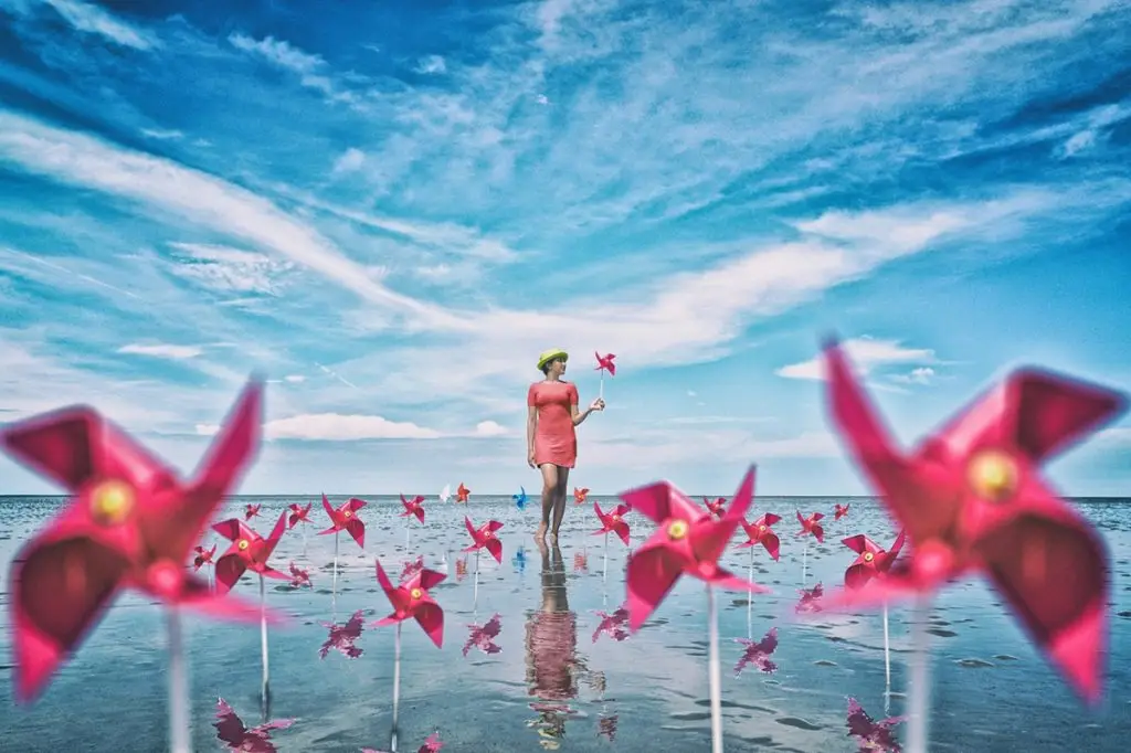 A woman in a coral dress holding a pinwheel on mirror-still shallow water under a wide blue sky, red pinwheels in the foreground