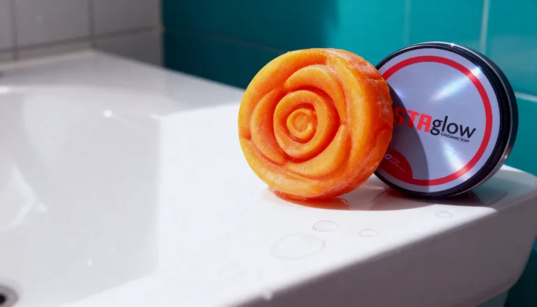 Orange rose-shaped soap leaning against an upright ASTAglow tin on a white sink with water drops, teal tiles behind.