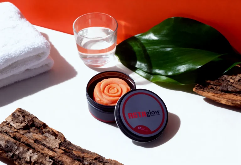 Open ASTAglow tin with rose soap, lid beside it, among towel, water glass, green leaf and bark on red-orange backdrop.