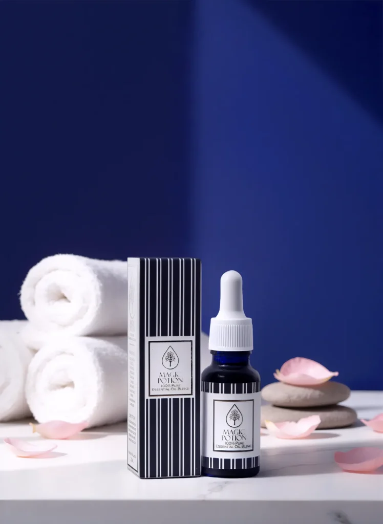 Magic Potion dropper bottle and striped box on white marble with rolled towels, spa stones and pink rose petals.