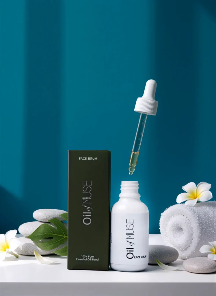 A dropper floats above an open white Oil of Muse bottle beside its green box, frangipani, stones and towel.
