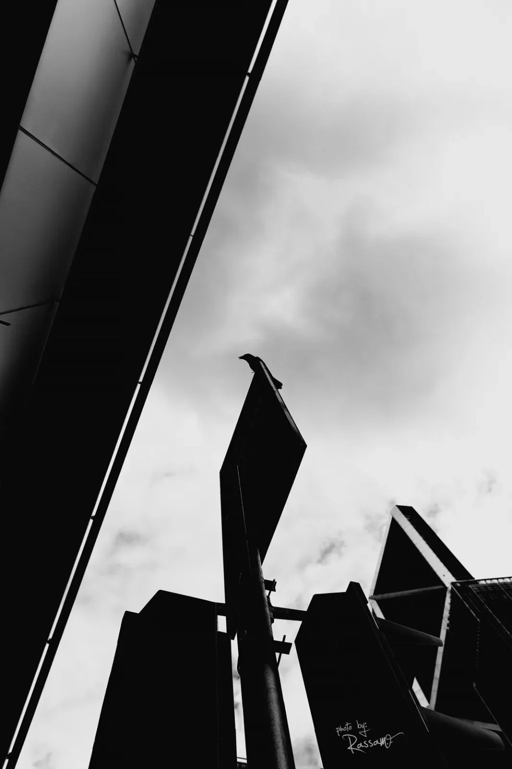 A crow perched on a tall sign between towers under a diagonal beam