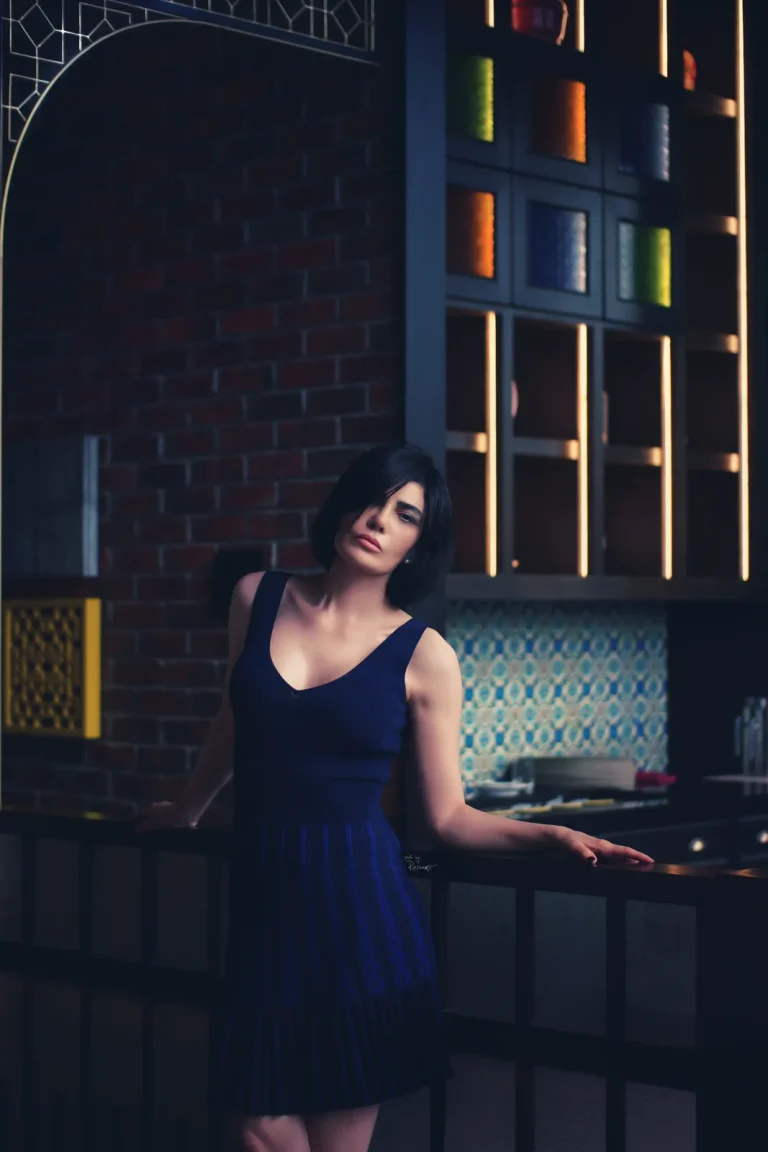 Woman in a navy knit dress leans on a dark counter, with brick wall, lit glass cabinets and tiles behind.