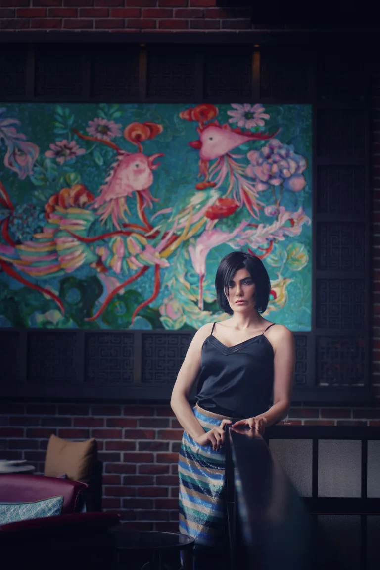 Woman in a black satin camisole and striped sequin skirt stands at a railing beneath a painting of pink birds.
