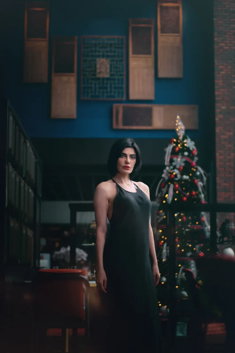 Woman in a black halter dress stands beside a lit Christmas tree, antique wooden panels on a blue wall above.