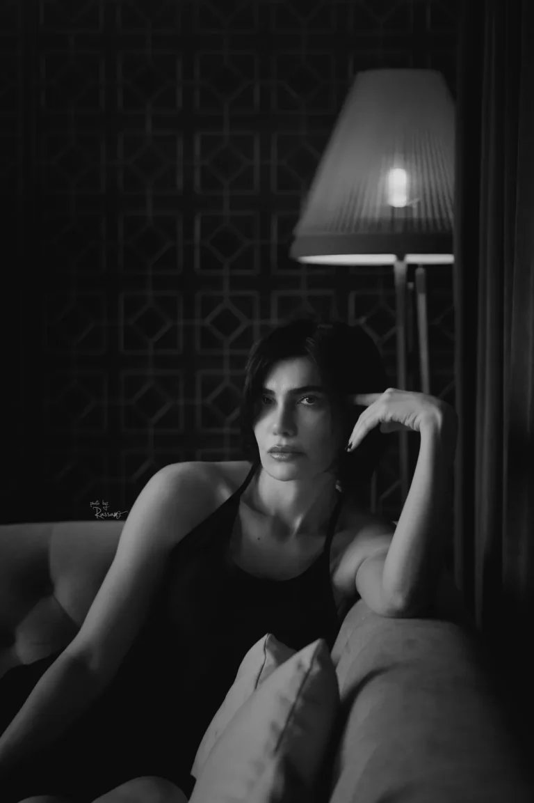 Black-and-white portrait of a woman on a sofa, finger at her temple, beside a glowing pleated lamp and lattice screen.