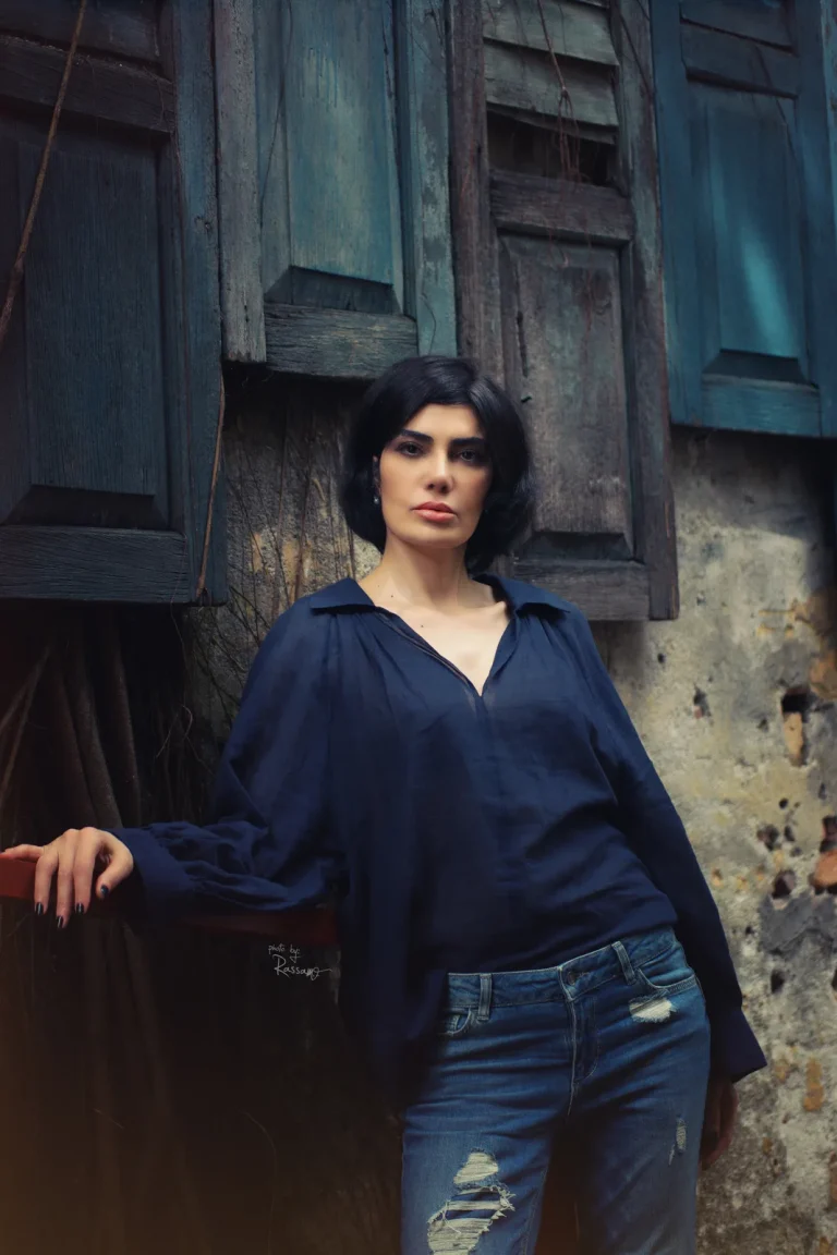 Woman in a sheer navy blouse and ripped jeans leans against a weathered wall with old teal wooden shutters.
