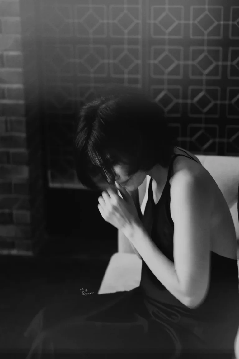 Black-and-white image of a woman with a black bob bowing her head, hands at her face, in soft hazy light.