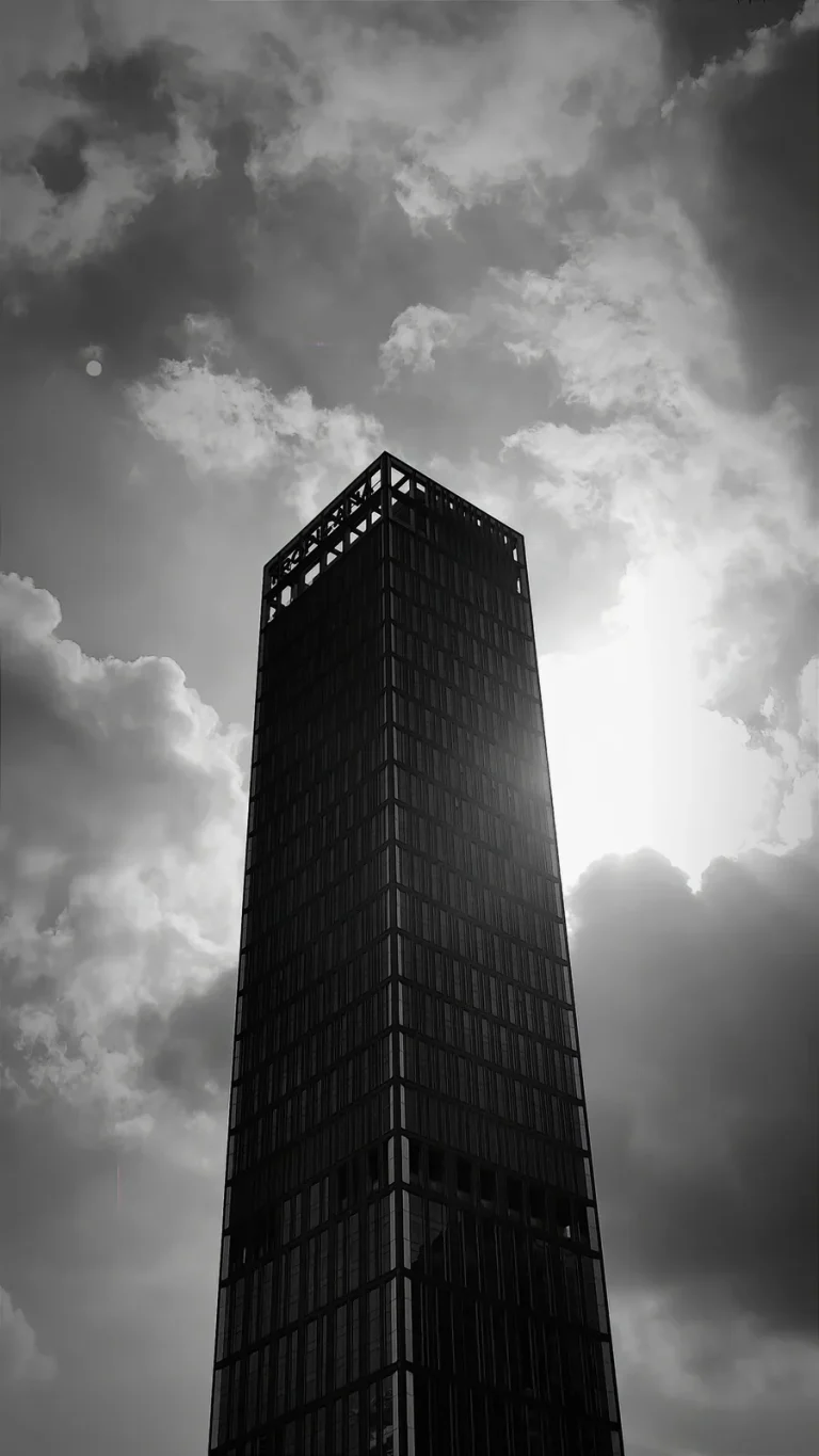 Black and white low-angle view of a dark rectangular skyscraper silhouetted against bright clouds and sun.