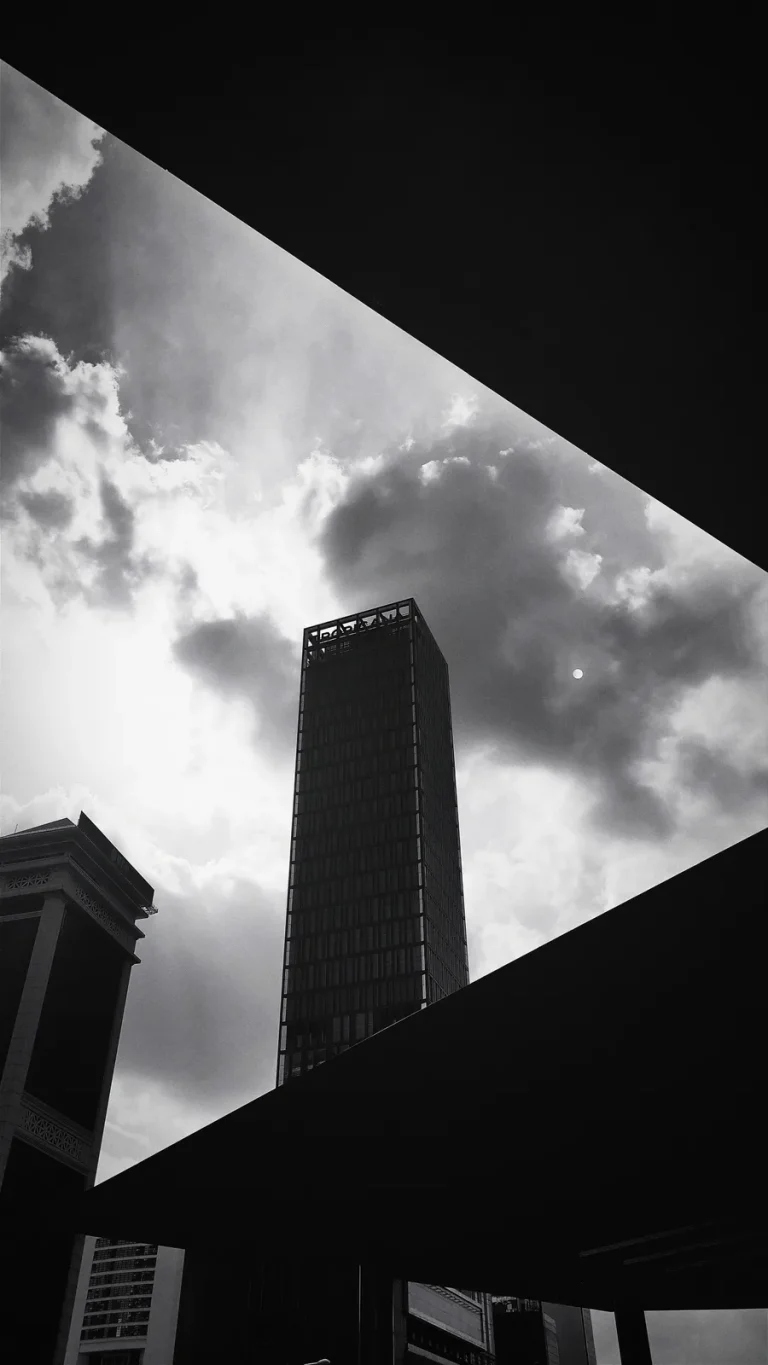 Black and white skyscraper framed by dark diagonal canopy edges under a bright, cloudy sky with the sun.