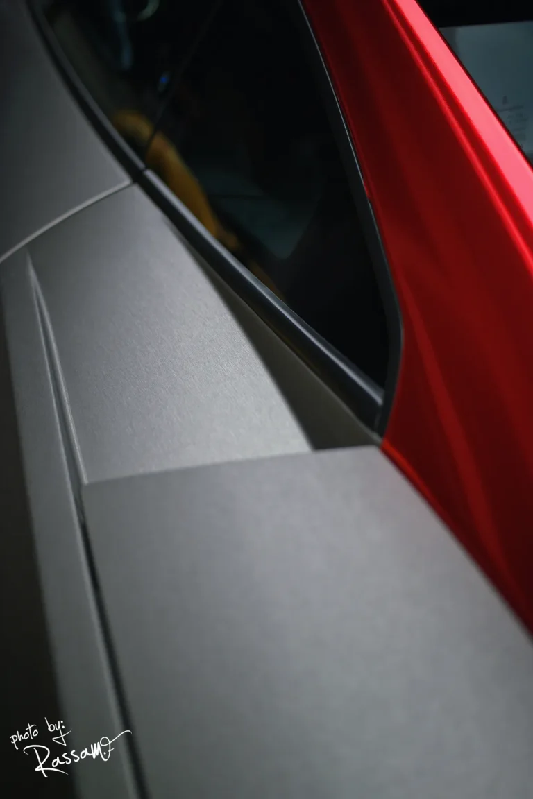 Abstract side detail of a Lamborghini where brushed grey panels meet a red-chrome pillar and dark tinted glass.