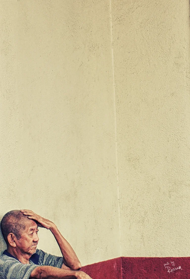 Elderly man resting a hand on his head against a cream wall above a red band