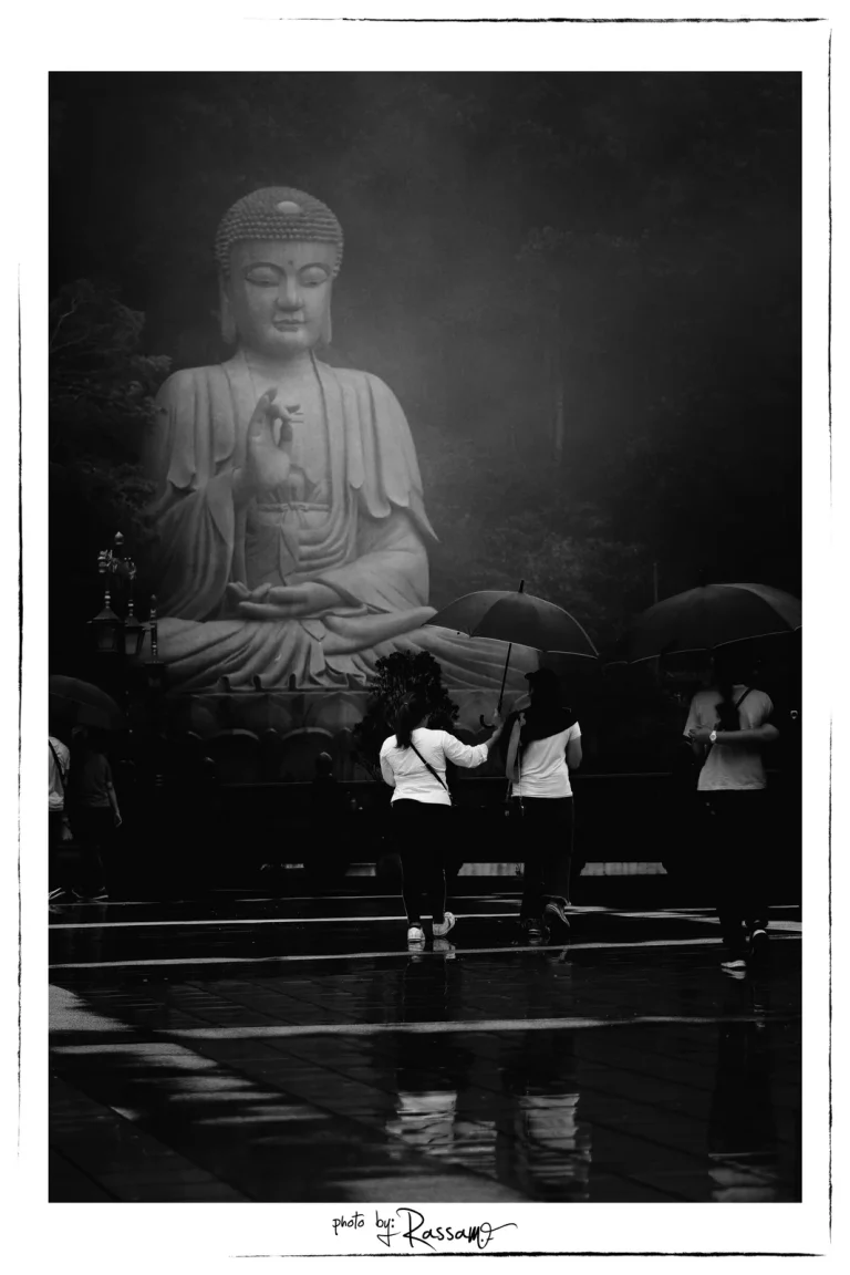 A giant seated Buddha statue above a wet courtyard where visitors walk with umbrellas, black and white