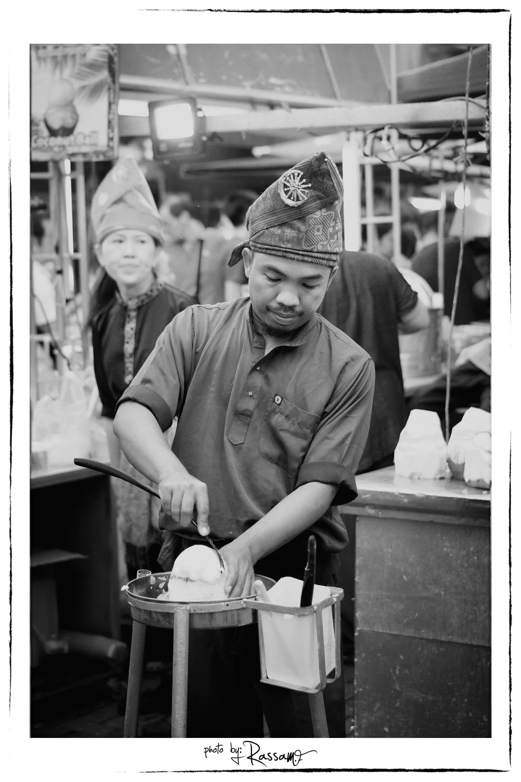 A man in traditional headwear opening a coconut at a market stall while a woman behind him smiles, black and white