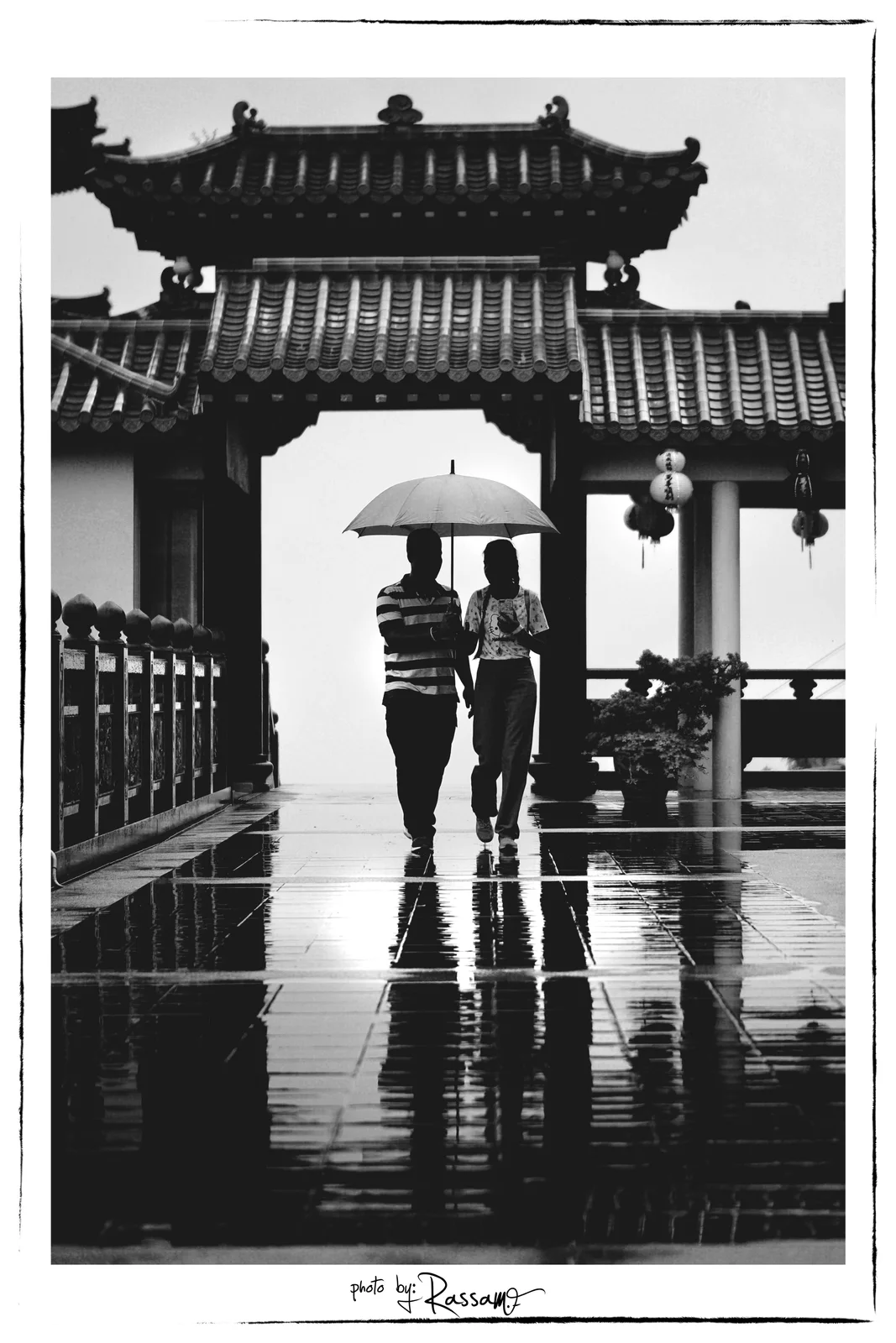 A couple sharing one umbrella walking through a temple gate, reflected in the wet floor, black and white