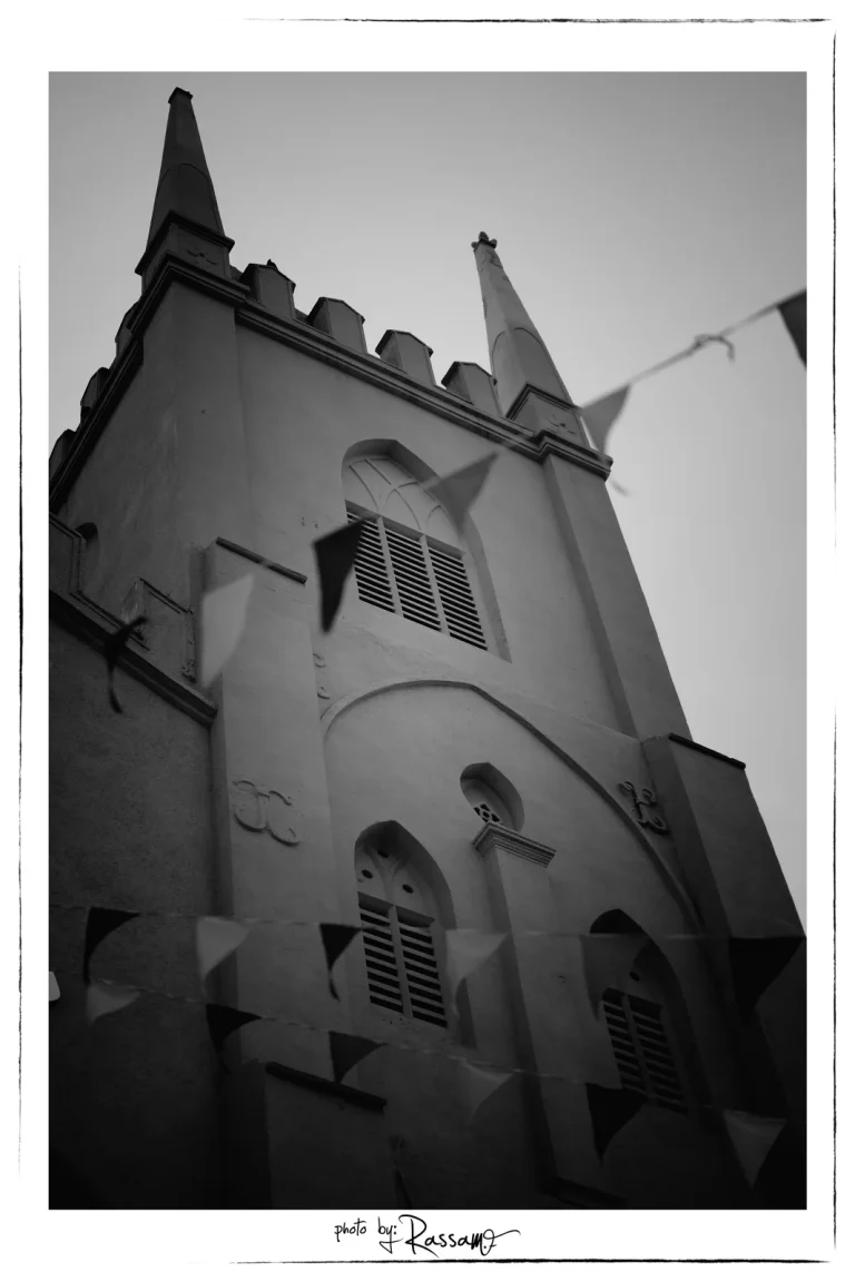 The church's side tower seen from below at an angle, with a string of festival flags across the frame, black and white