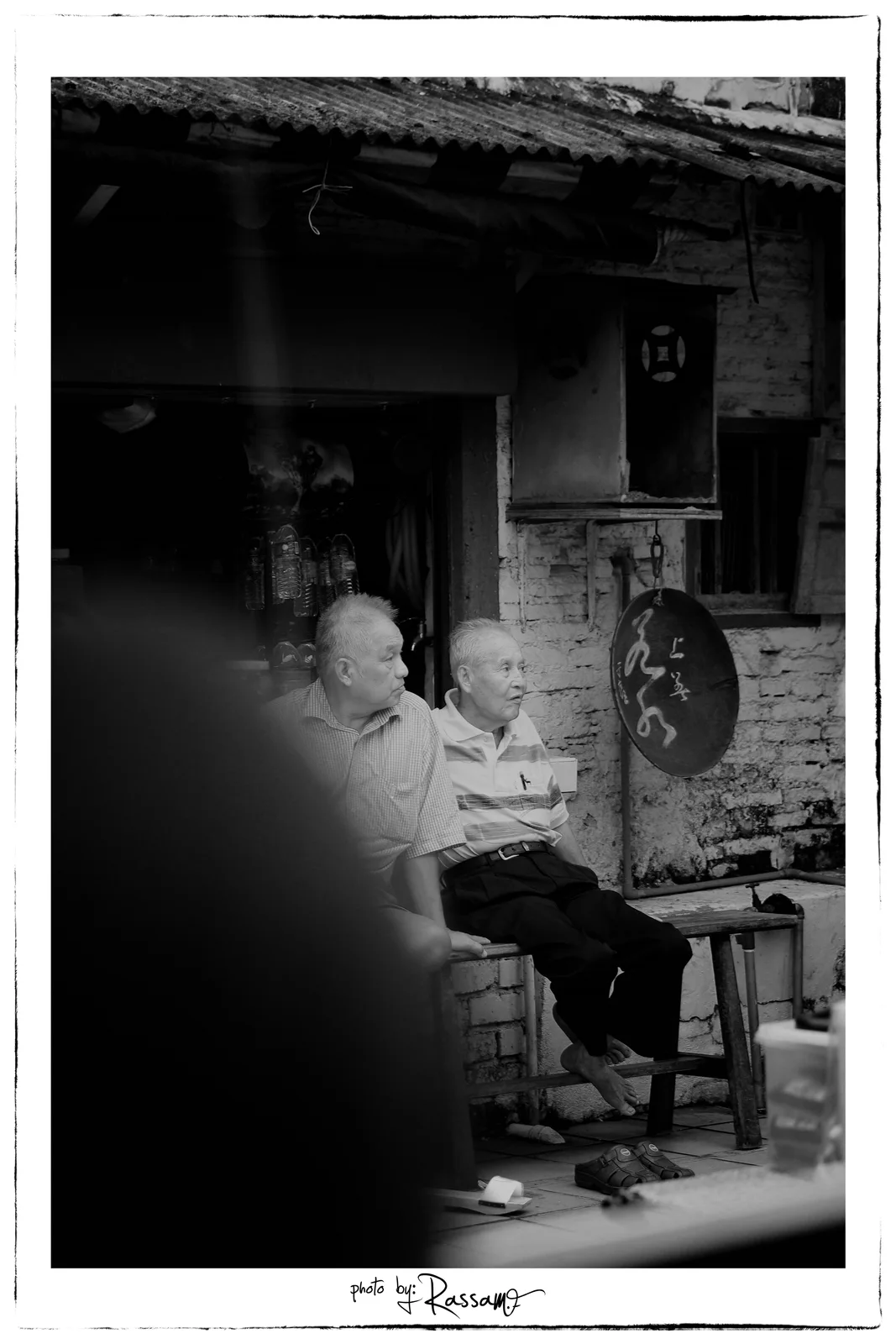 Two older men sitting on a bench outside an old shop, a blurred dark shape in the foreground, black and white